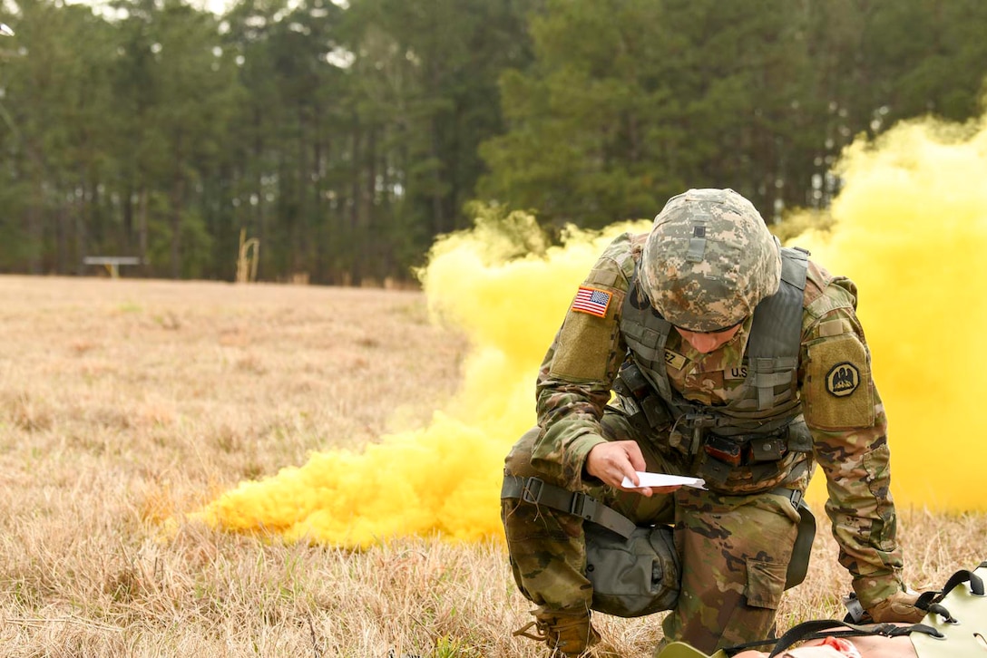 A guardsman kneels in a field and looks at a piece of paper as yellow smoke billows behind him.