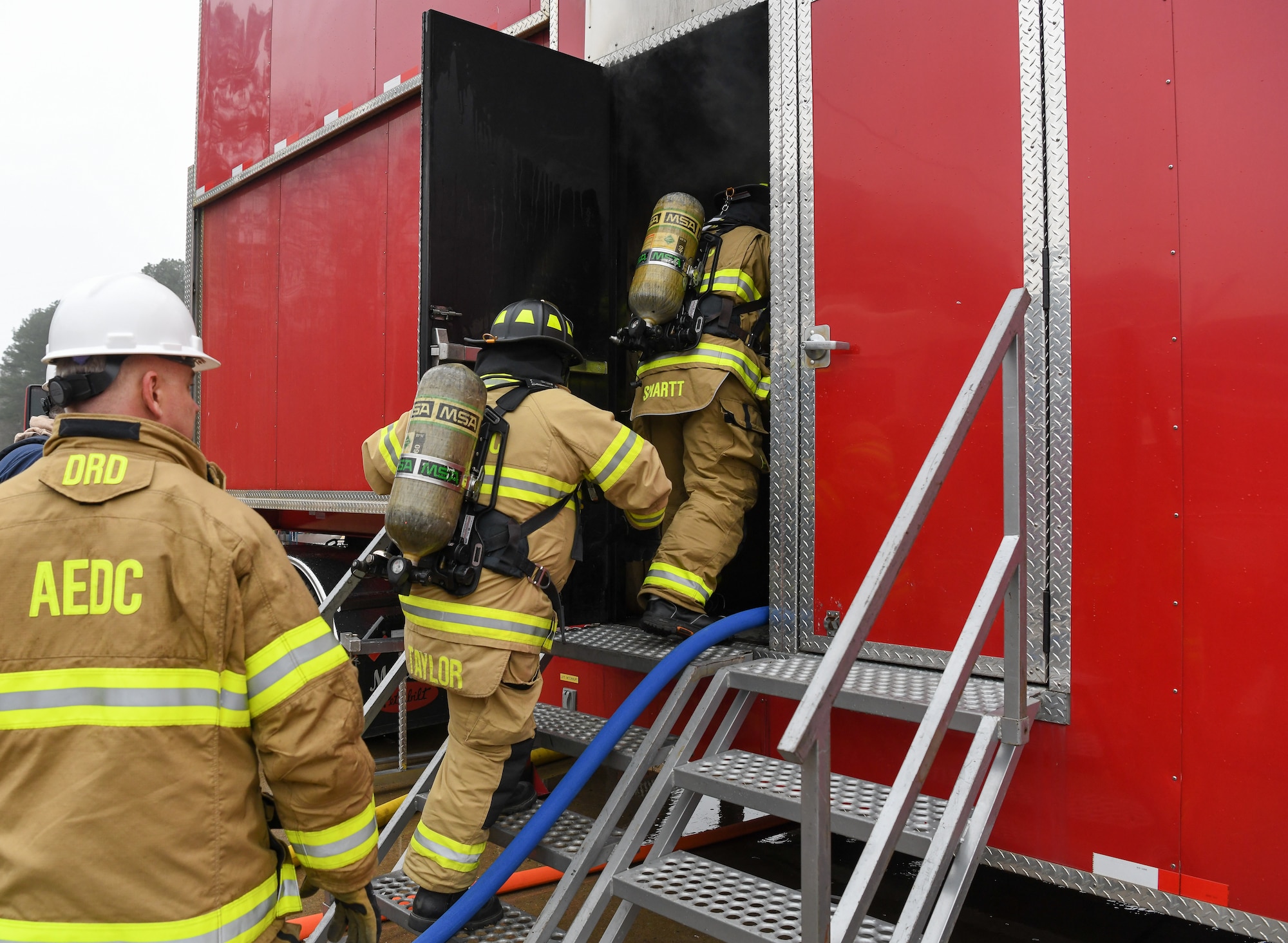 Two firefighters in turnout gear entering training trailer as another watches