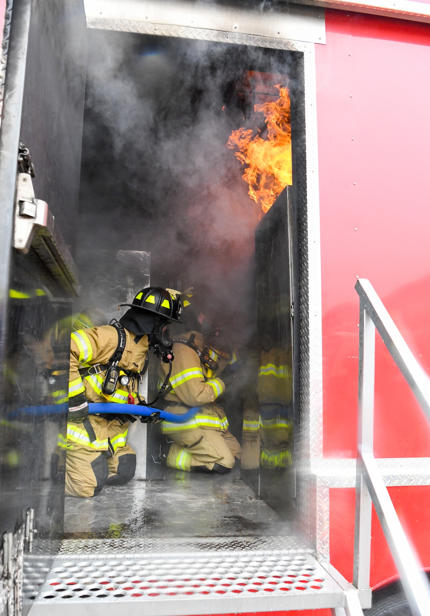 Firefighters in turnout gear on knees inside trailer with flames and simulated smoke