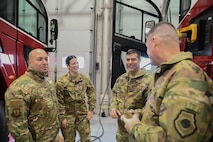 Gen Gebara and Chief Master Sgt. Cenov talk with Col. Walters and an Airman in between two fire trucks inside the fire department.