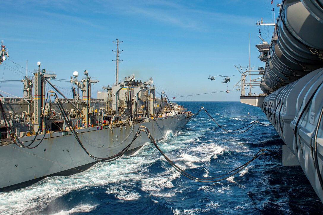 Two ships transit a body of water during a replenishment as two helicopters fly nearby.