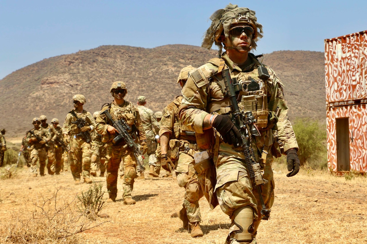 Many soldiers walk through a dry, rural area.