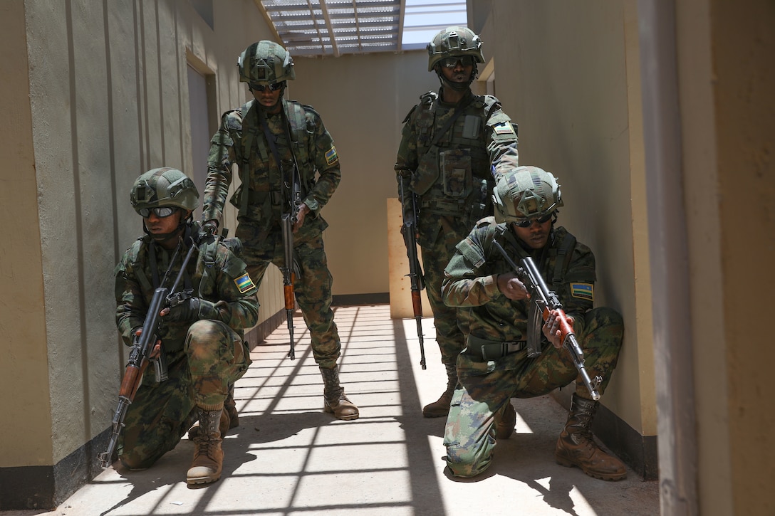 Two soldiers hold weapons as they squat next to one another in a narrow space. Behind them, two soldiers hold weapons.