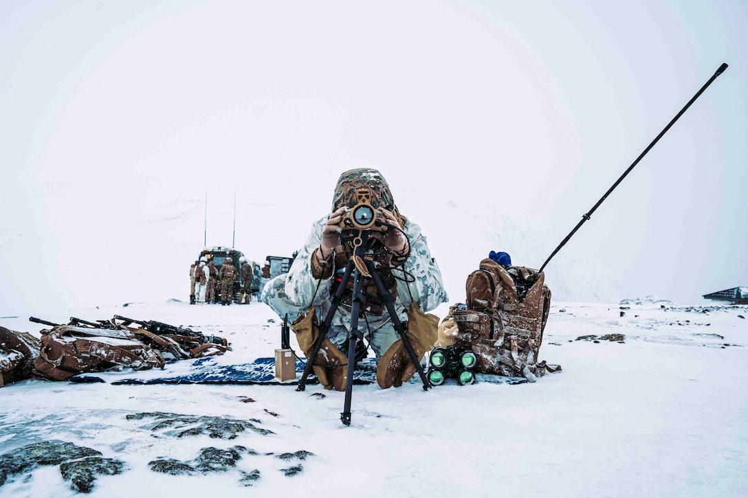 A Marine looks through binoculars while kneeling in snow.