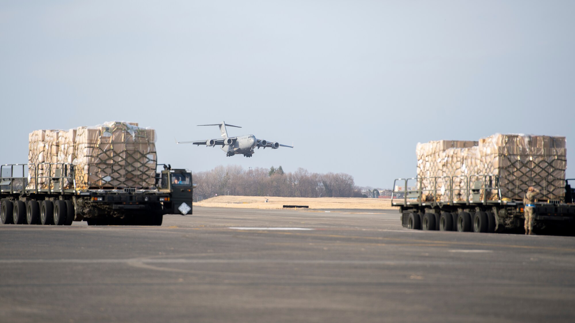 A large military cargo aircraft comes in for a landing with two heavily loaded cargo trucks waiting in the foreground.