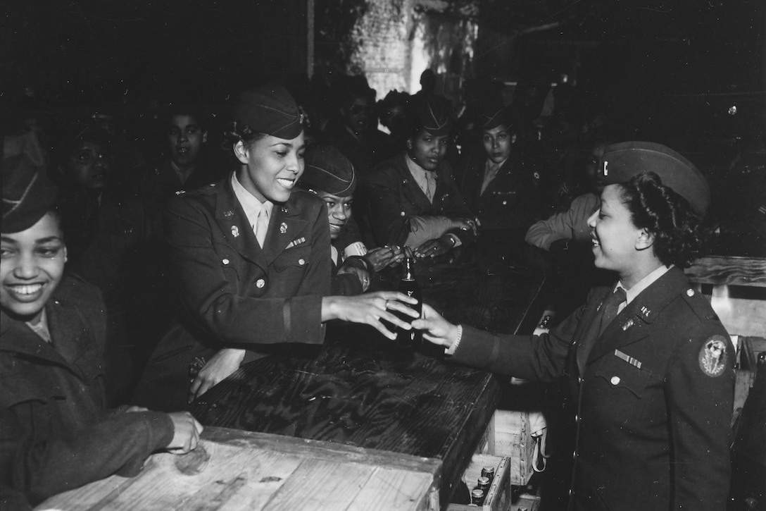 A woman behind a crowded bar serves a woman a bottle of soda.