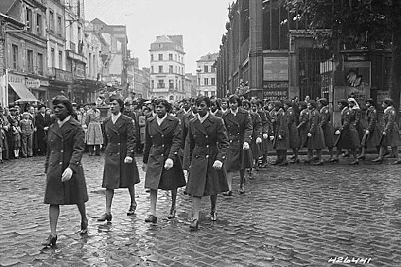 Women in uniform march in formation across a brick-paved street.