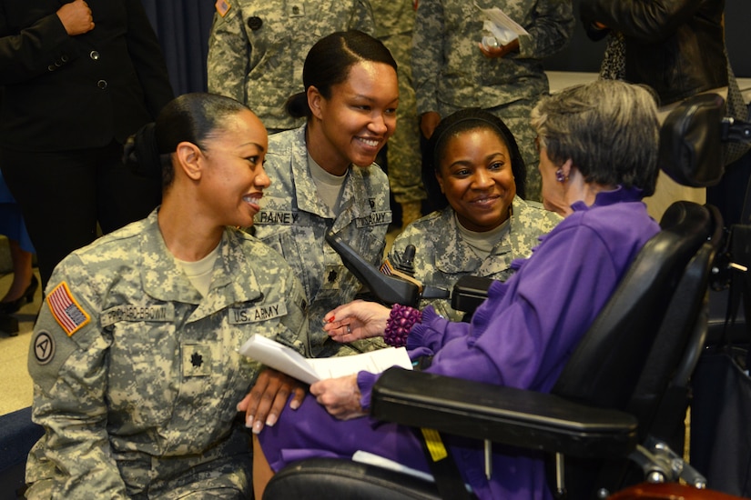 Three female soldiers smile at an elderly woman in a wheelchair.