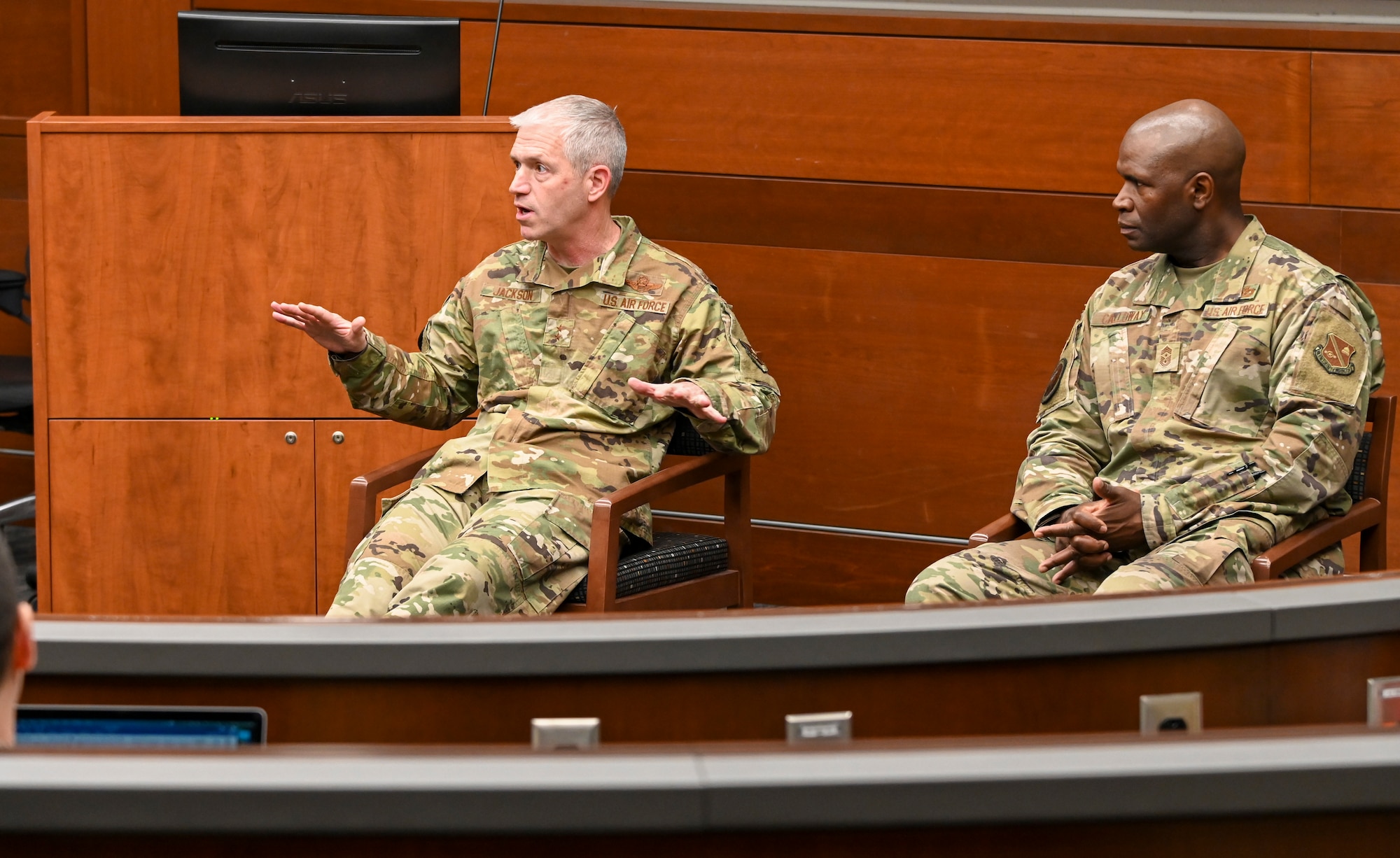 Two men are seated in classroom, speaking to group (not shown).