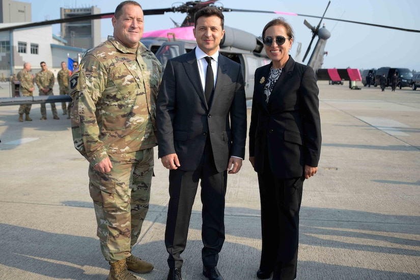 Two men and a woman pose for a photo. A military aircraft is in the background.
