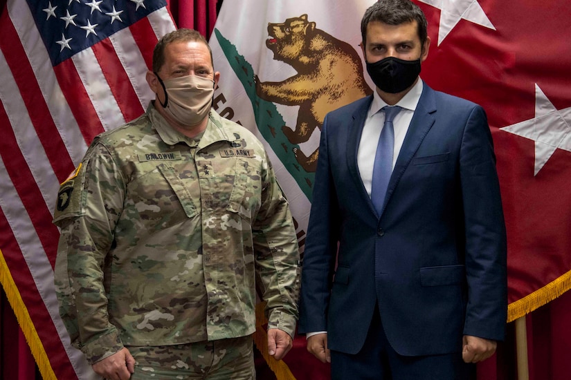 A service member and civilian stand in front of flags.