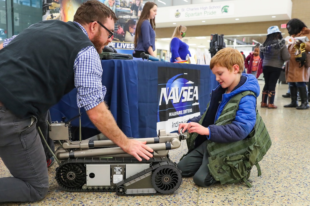 An NSWC IHD employee is kneeling on the floor describing an explosive ordnance disposal robot to a young boy.