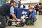 An NSWC IHD employee is kneeling on the floor describing an explosive ordnance disposal robot to a young boy.