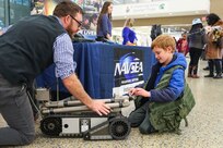 An NSWC IHD employee is kneeling on the floor describing an explosive ordnance disposal robot to a young boy.