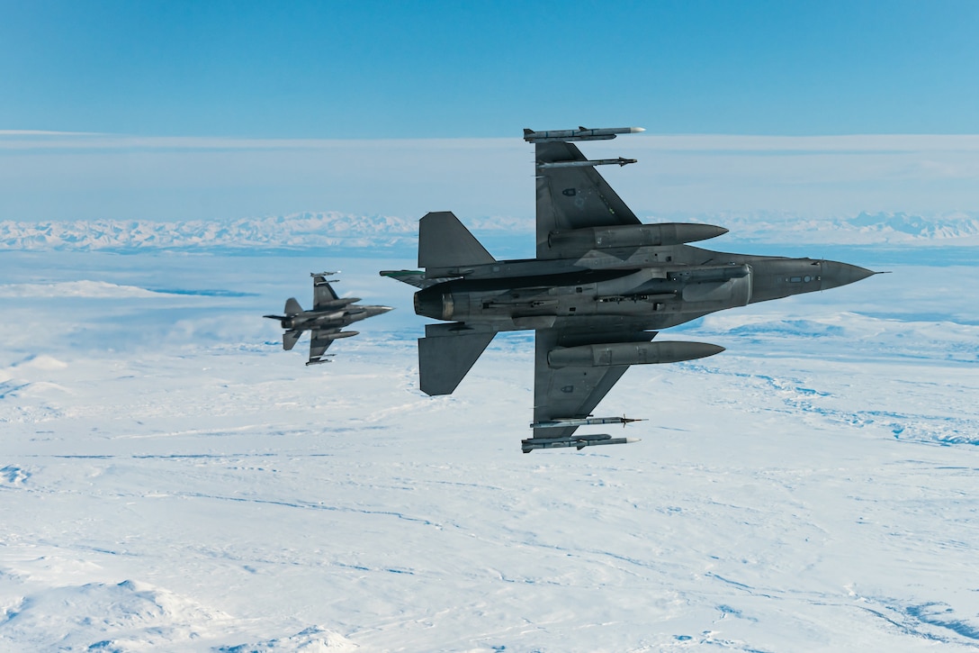 Two military jets fly above icy terrain.