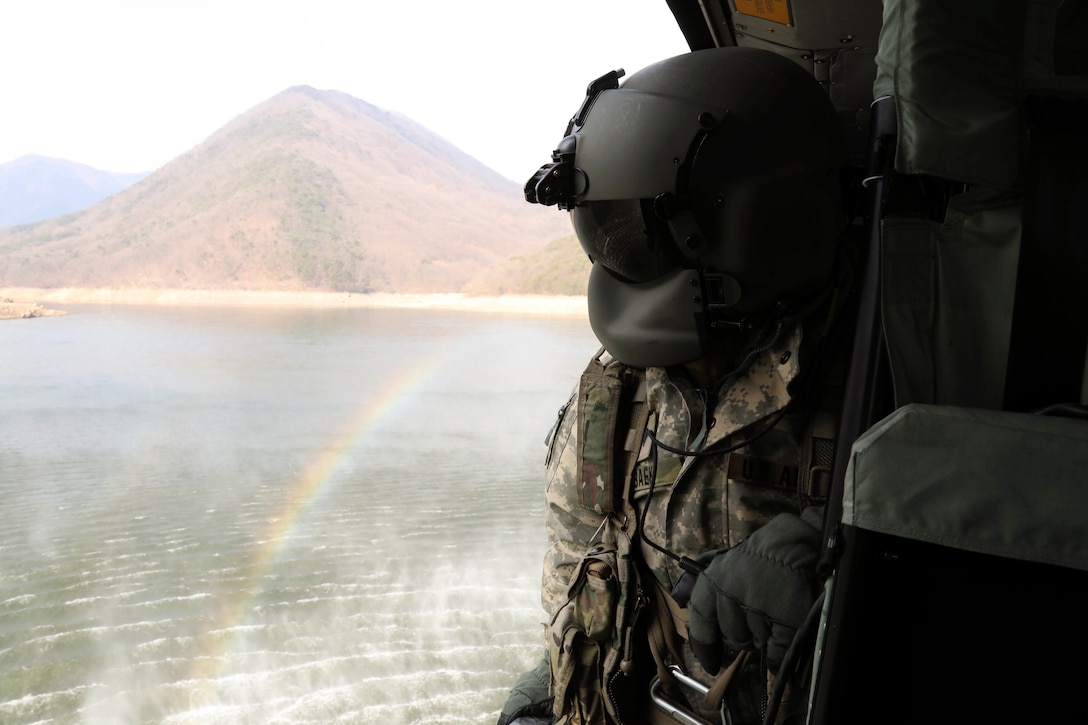 A solider looks out of an airborne aircraft as it flies over a body of water near a rainbow and mountains.