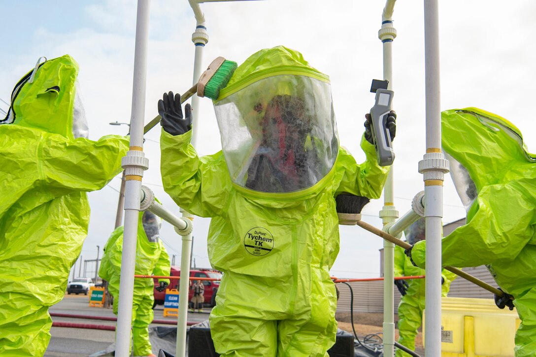 Airmen wearing neon hazard suits use brushes to scrub a fellow airman after hazardous materials decontamination training.