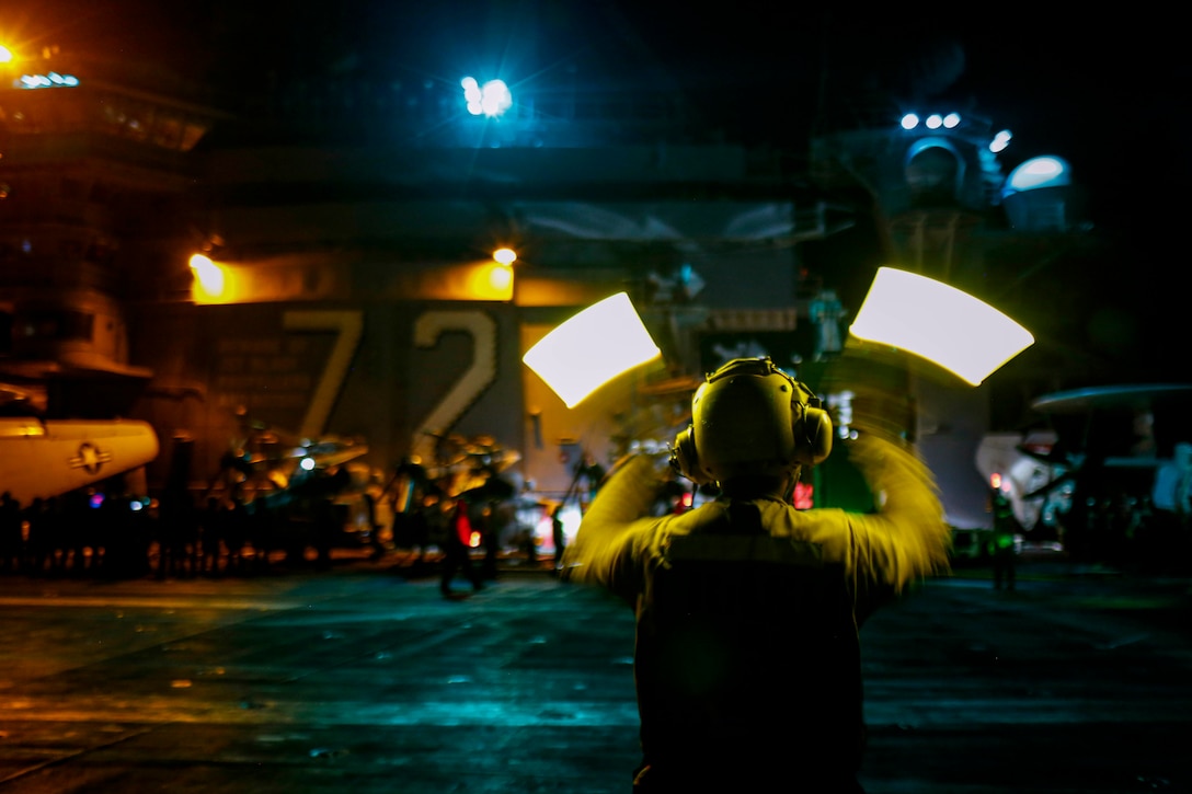 A sailor waves batons to direct a crane aboard a ship at sea in the dark illuminated by neon lights.