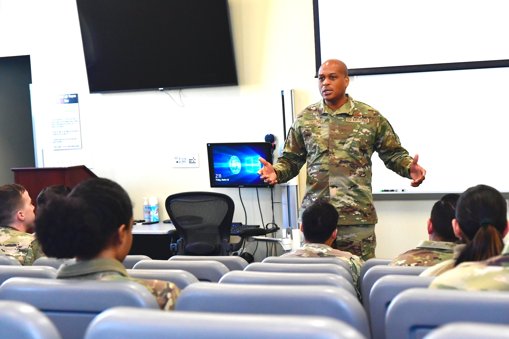 Brig. Gen. Roy Collins gestures with open arms to Defenders seated in a small auditorium.