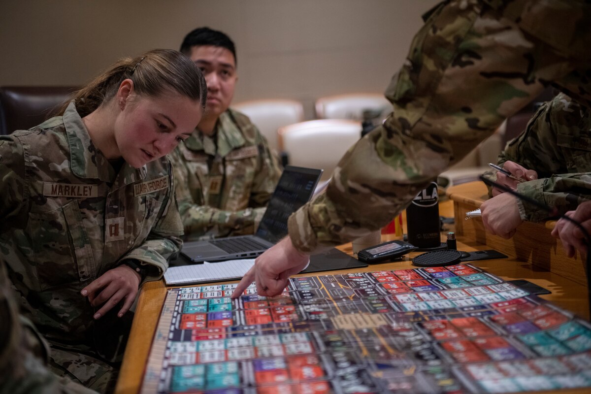 an arm entering the framefrom the top right points to a square on a board game while a participant looks at it