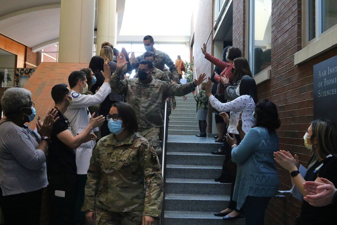 Airmen wearing face masks walk past university medical staff members wearing face masks while they clap and say goodbye.