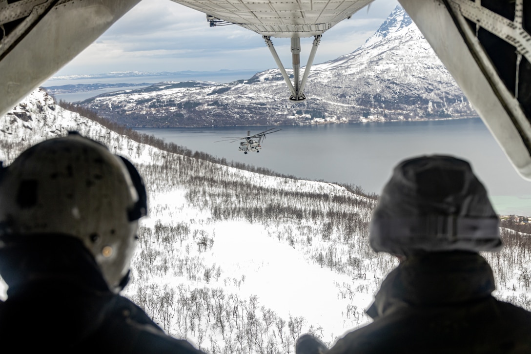 Two service members look at the snowy view from the back of a helicopter.