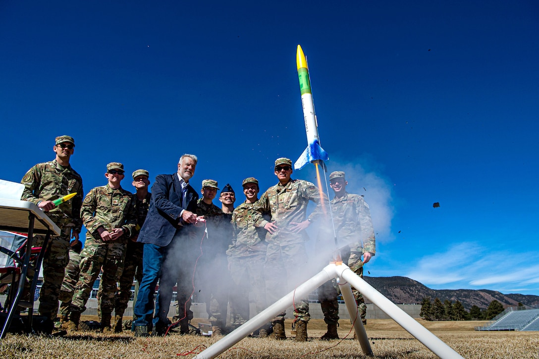 A group of service members watch as a man launches a small rocket.