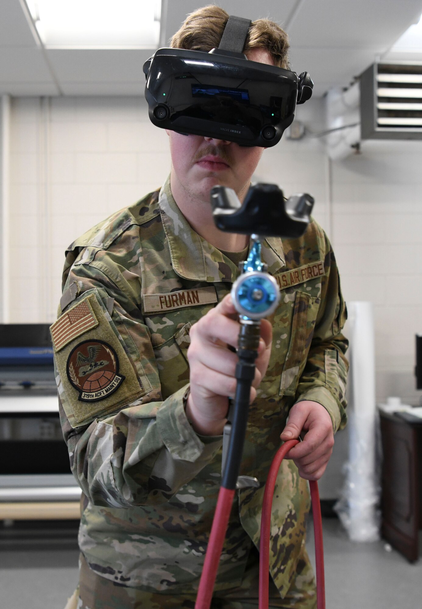 An Airman stands holding a paint gun that is connected to a training simulator.