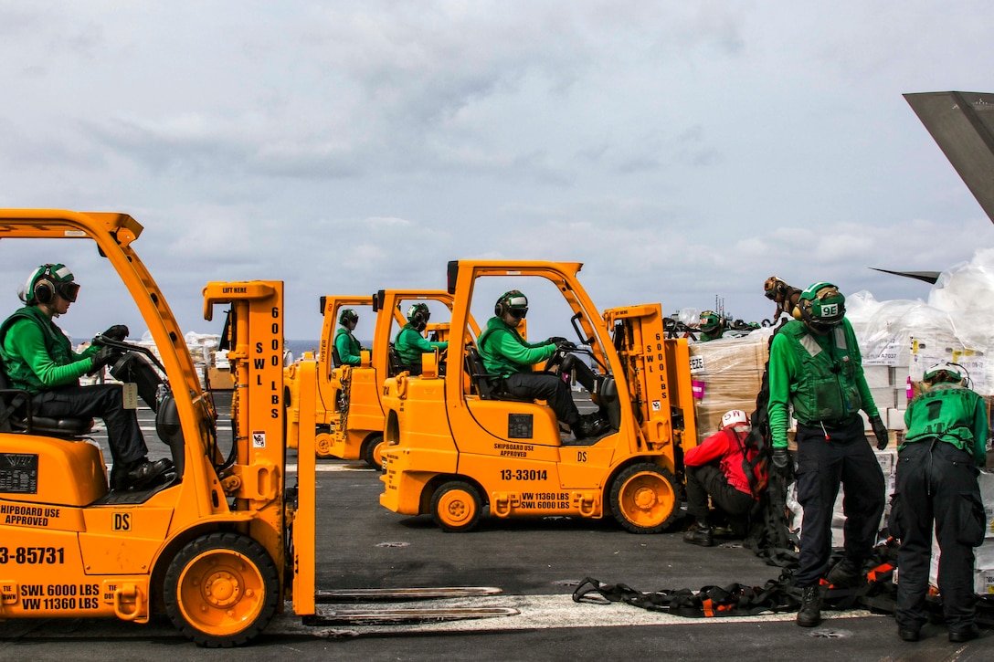 A group of sailors driving forklifts prepare to move pallets of supplies.