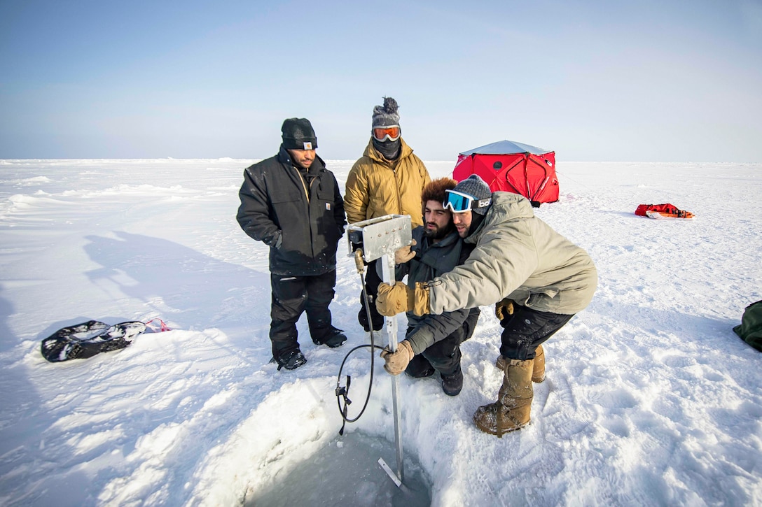 Personnel with Naval Undersea Warfare Center Newport and Arctic Submarine Laborator look at the monitor of an underwater camera while standing on the snow.