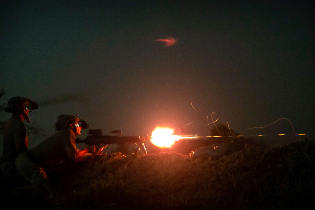 A solider fires a weapon at night while a fellow solider watches.