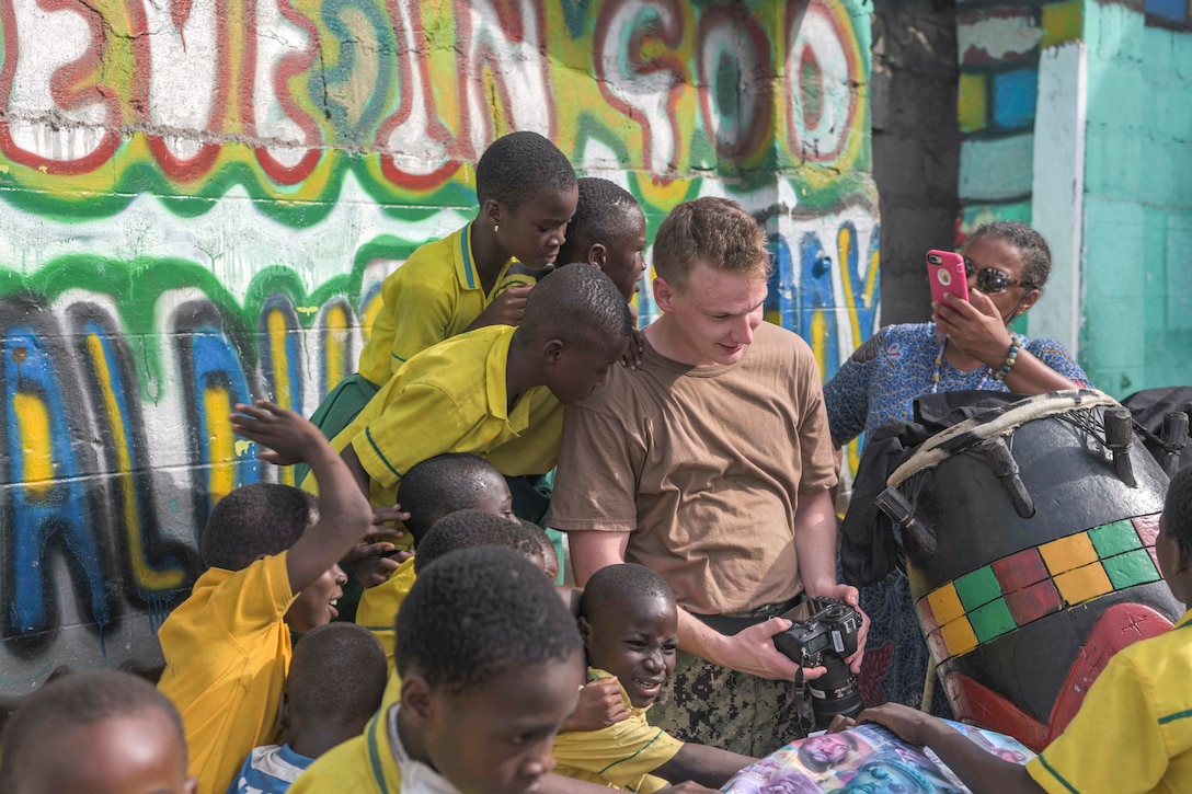 A sailor holds a camera and children gather around him and look at the screen.