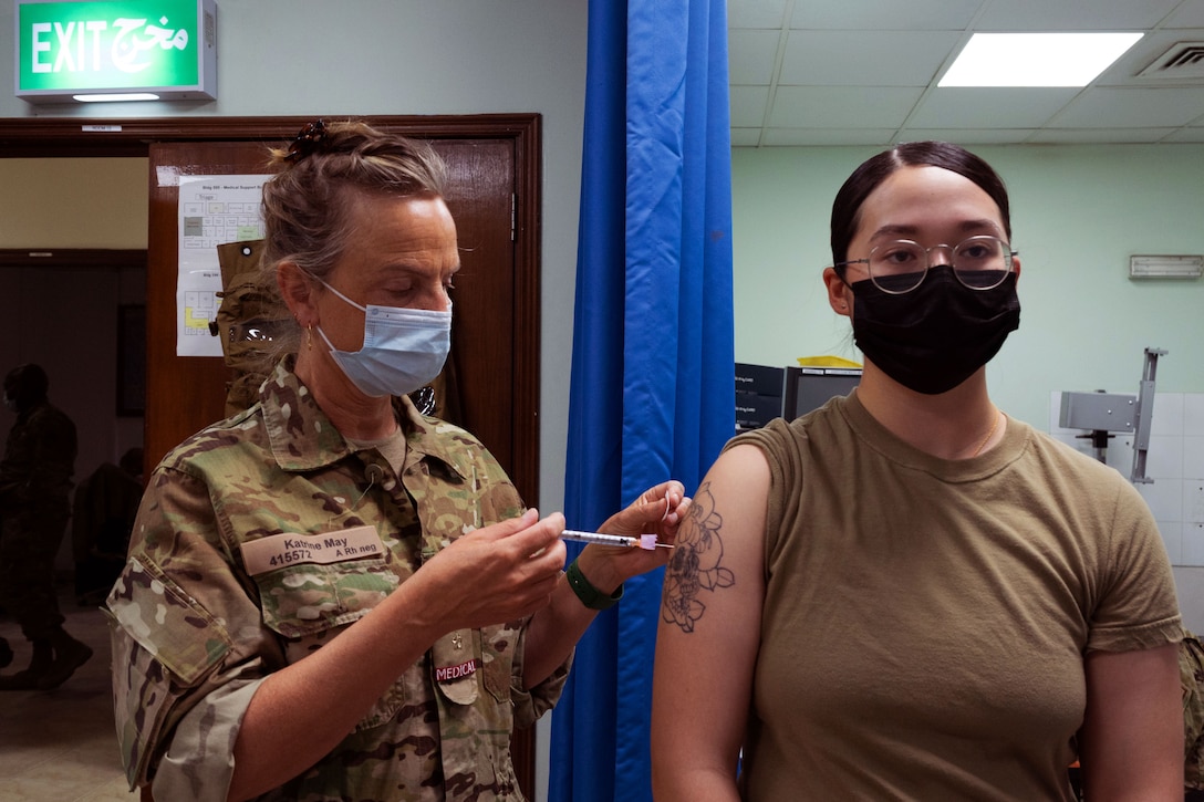 A service member gives an injection to a woman; both are wearing face masks.