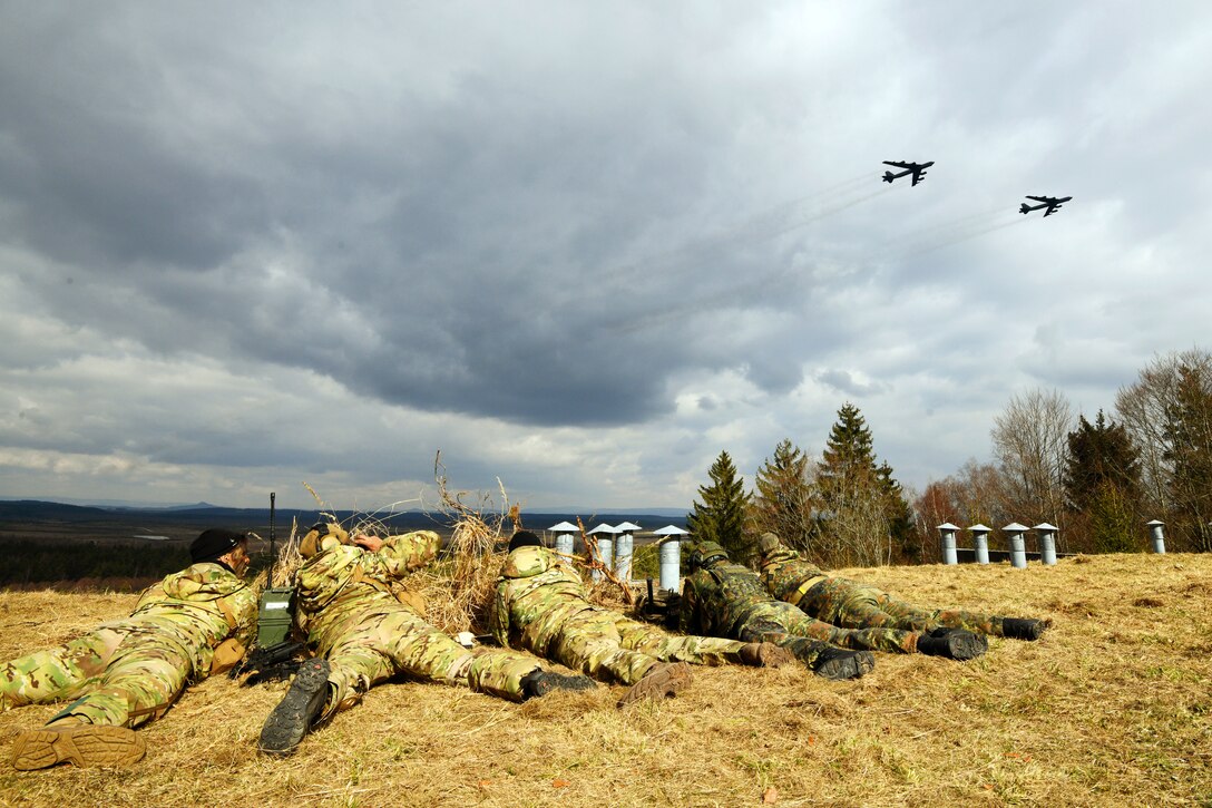 Five service members lie on their stomachs out in the field as two aircraft fly overhead.