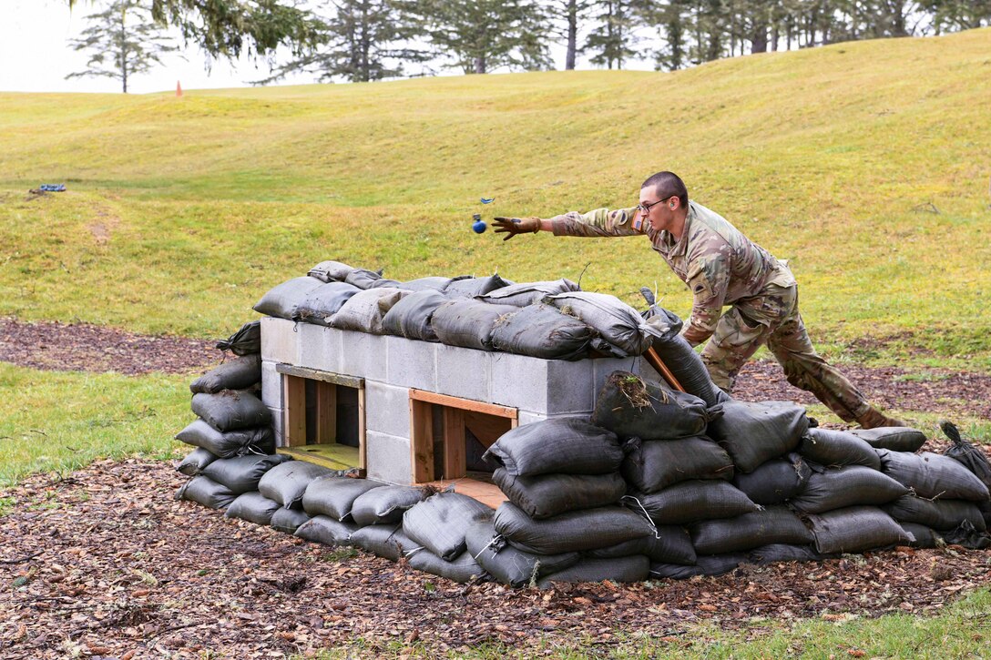 A National Guardsman standing behind a manmade barrier throws a mock grenade during training.