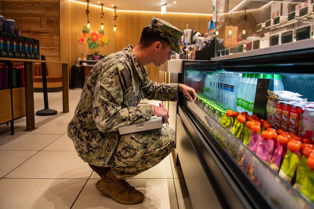 A sailor kneels next to a refridgerated beverage aisle .