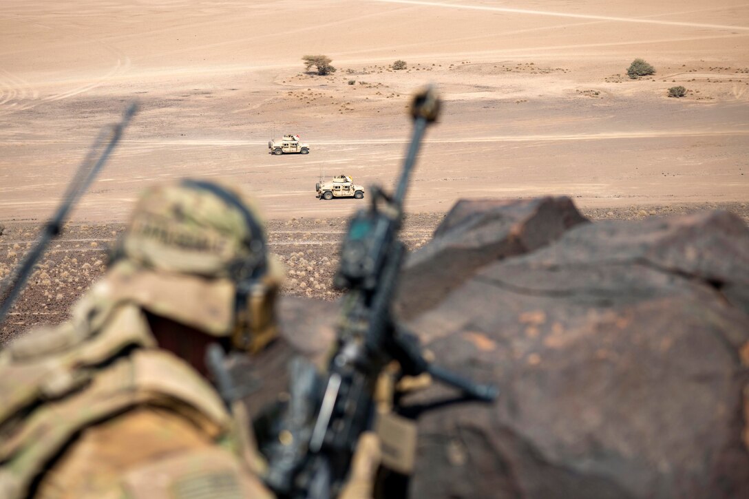 Soldiers holding weapons look at vehicles drive on a dirt road.