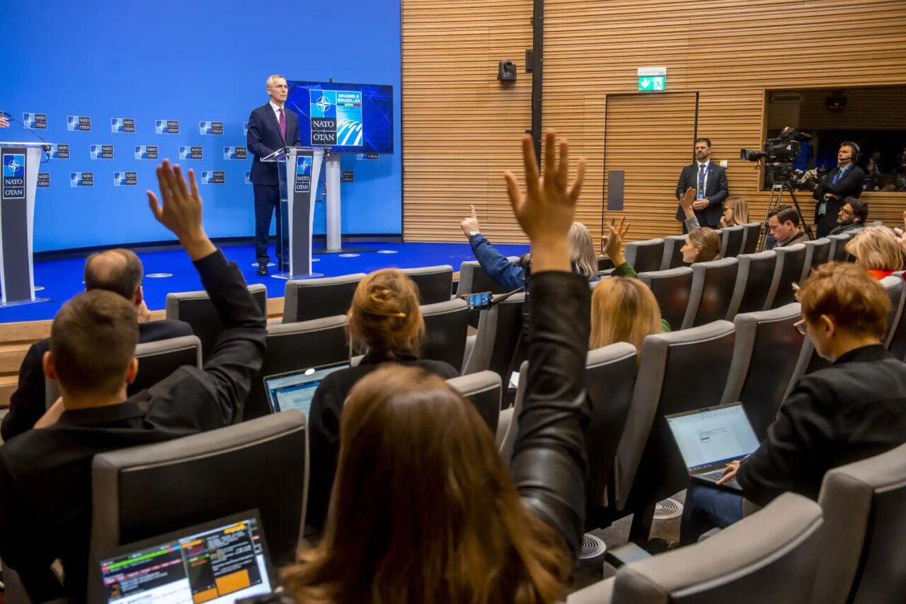 A man stands at a lectern; people in the audience raise their hands.