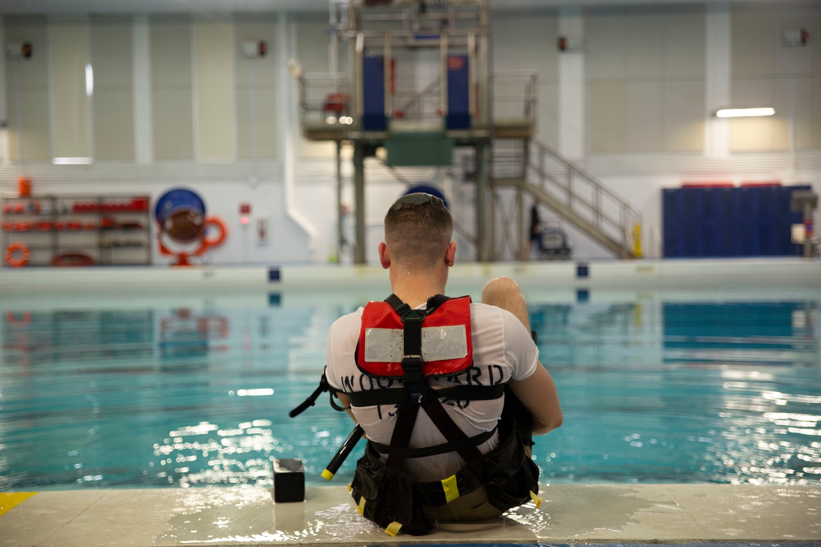 A prospective Coast Guard rescue swimmer sits at the edge of a training pool with their back to the camera