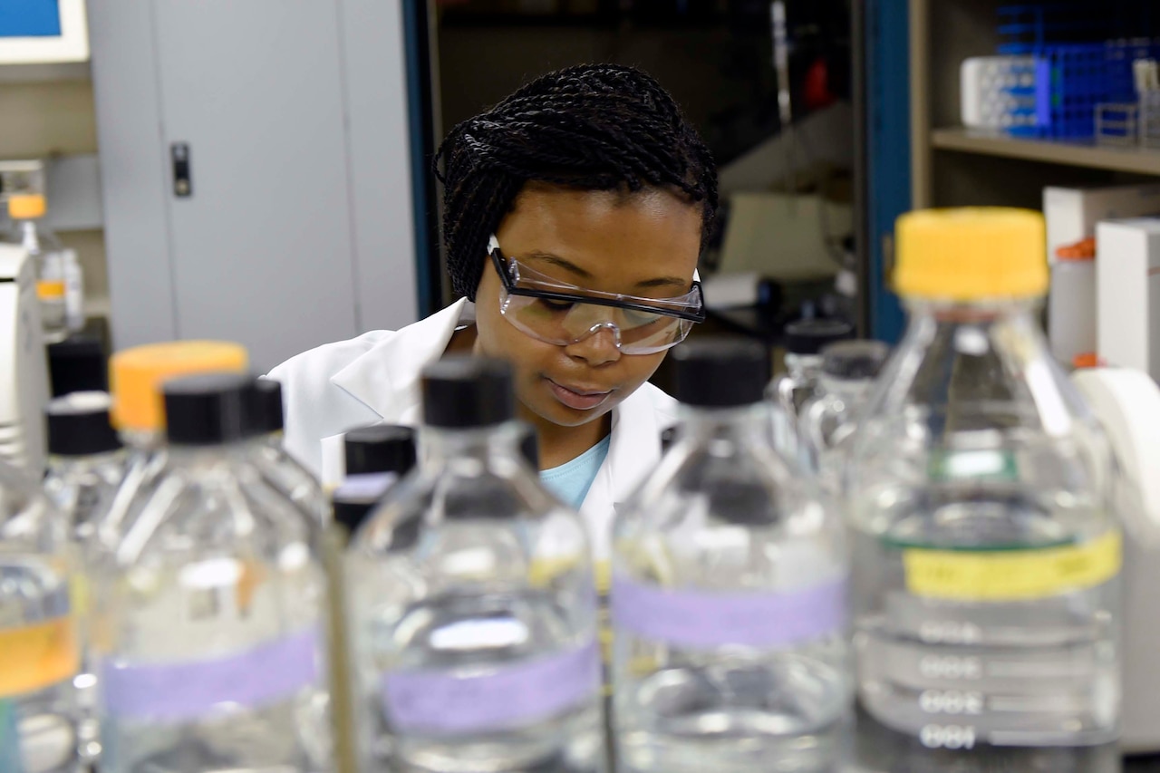 A woman in a lab coat stands behind an array of bottles.