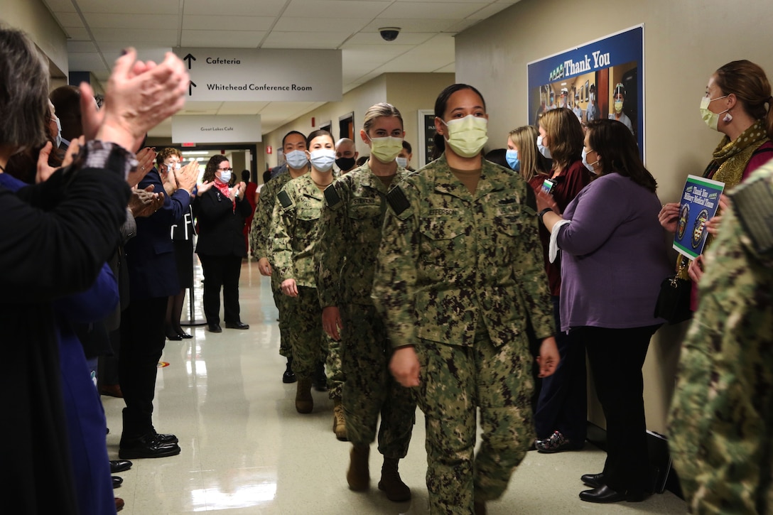 People wearing face masks stand along both sides of a corridor and clap as service members, who are wearing face masks, walk between them.