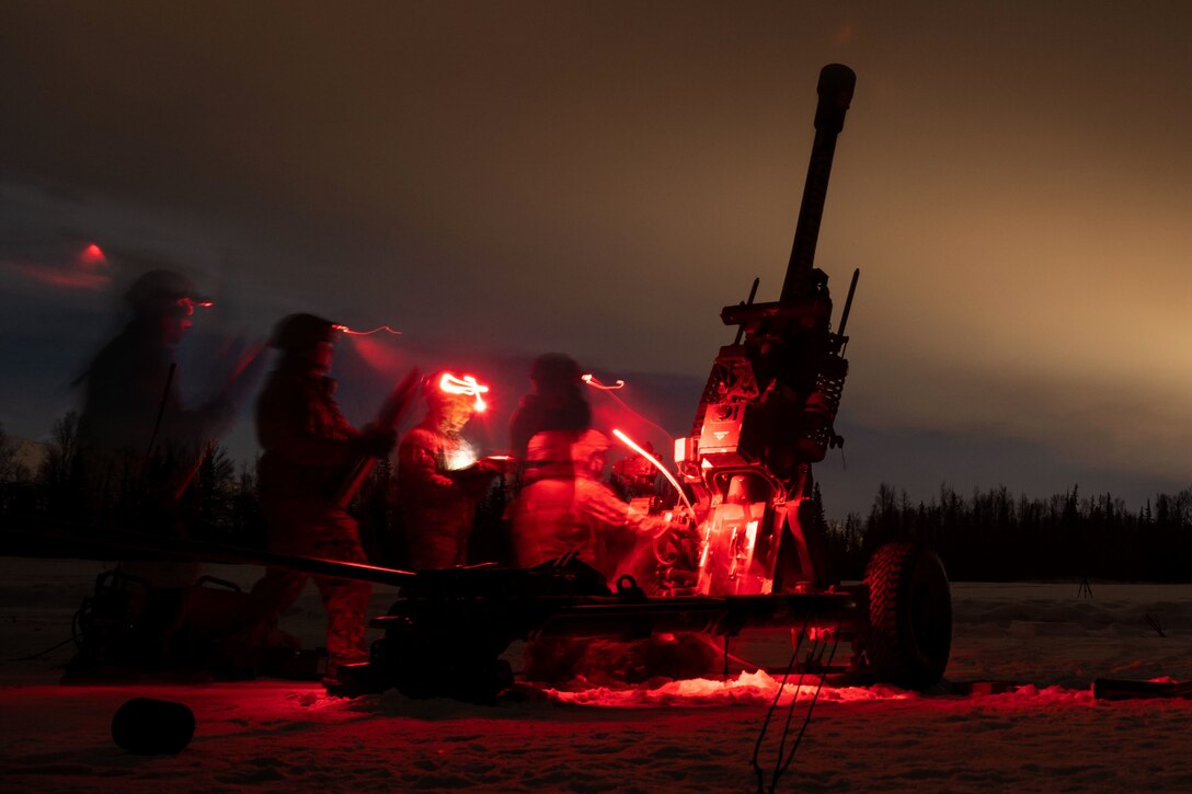 Several soldiers bathed in red light stand behind a howitzer at night.