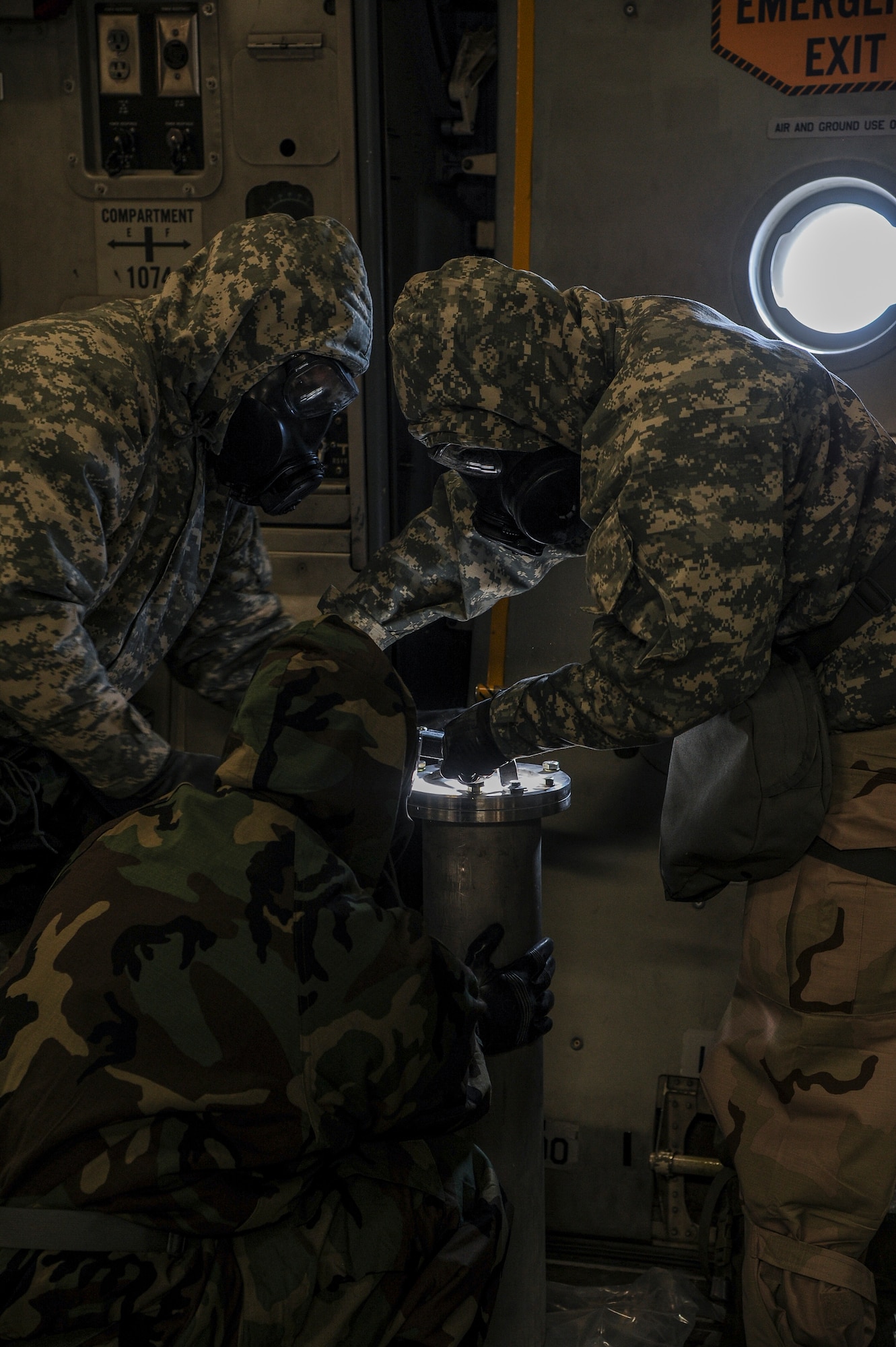 Soldiers in CBRN gear look at multiple round container on aircraft.
