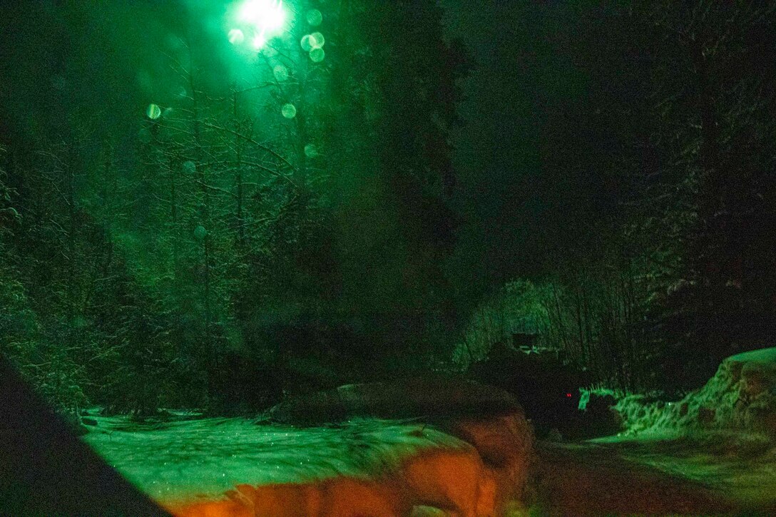 A military vehicle illuminated by green light drives down a road.