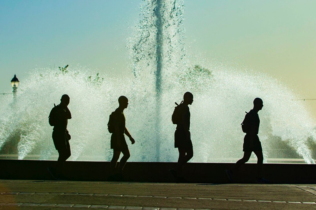 Four Marines shown in silhouette walk in front of a water fountain.