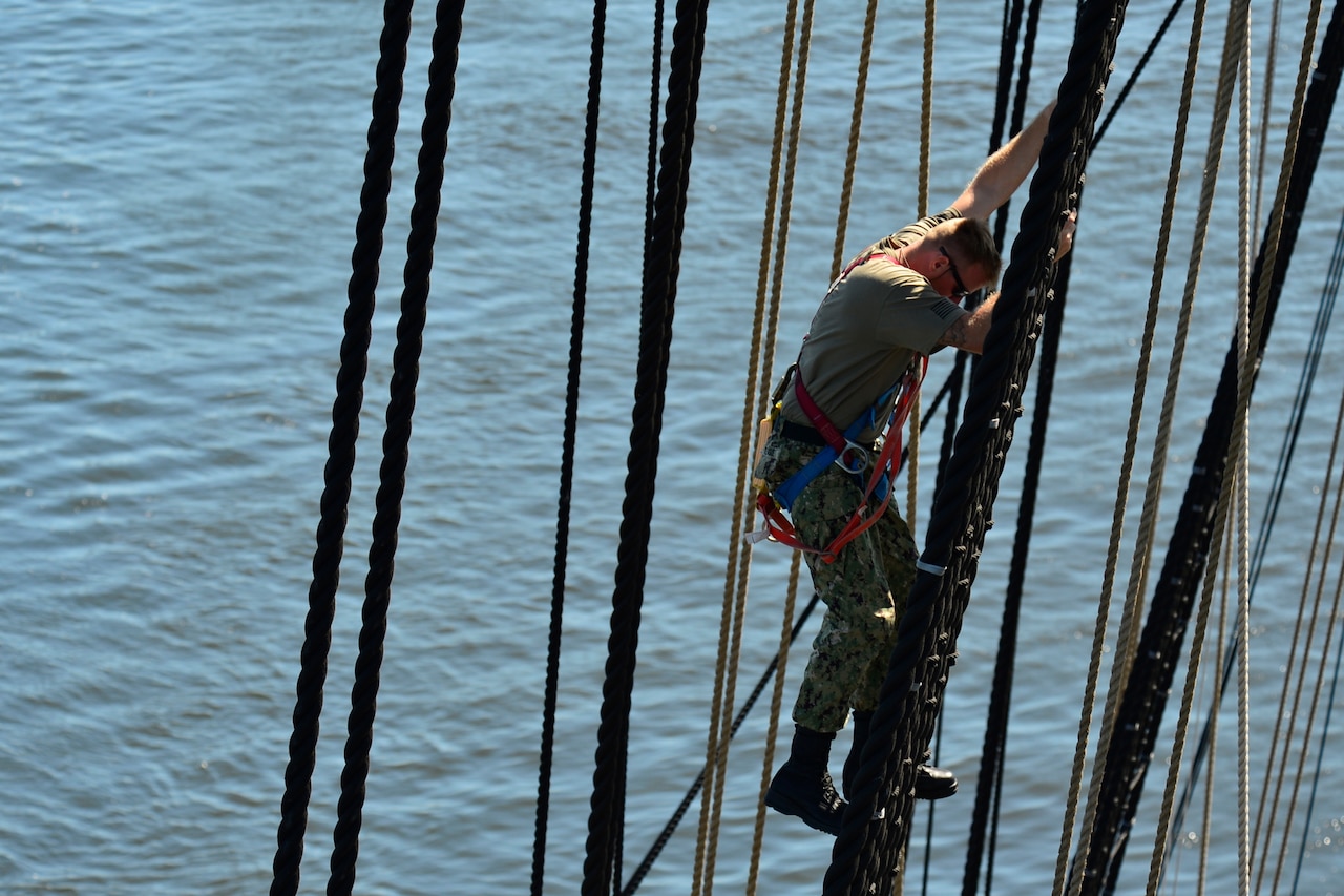 A man climbs the rigging of a large ship’s mast.