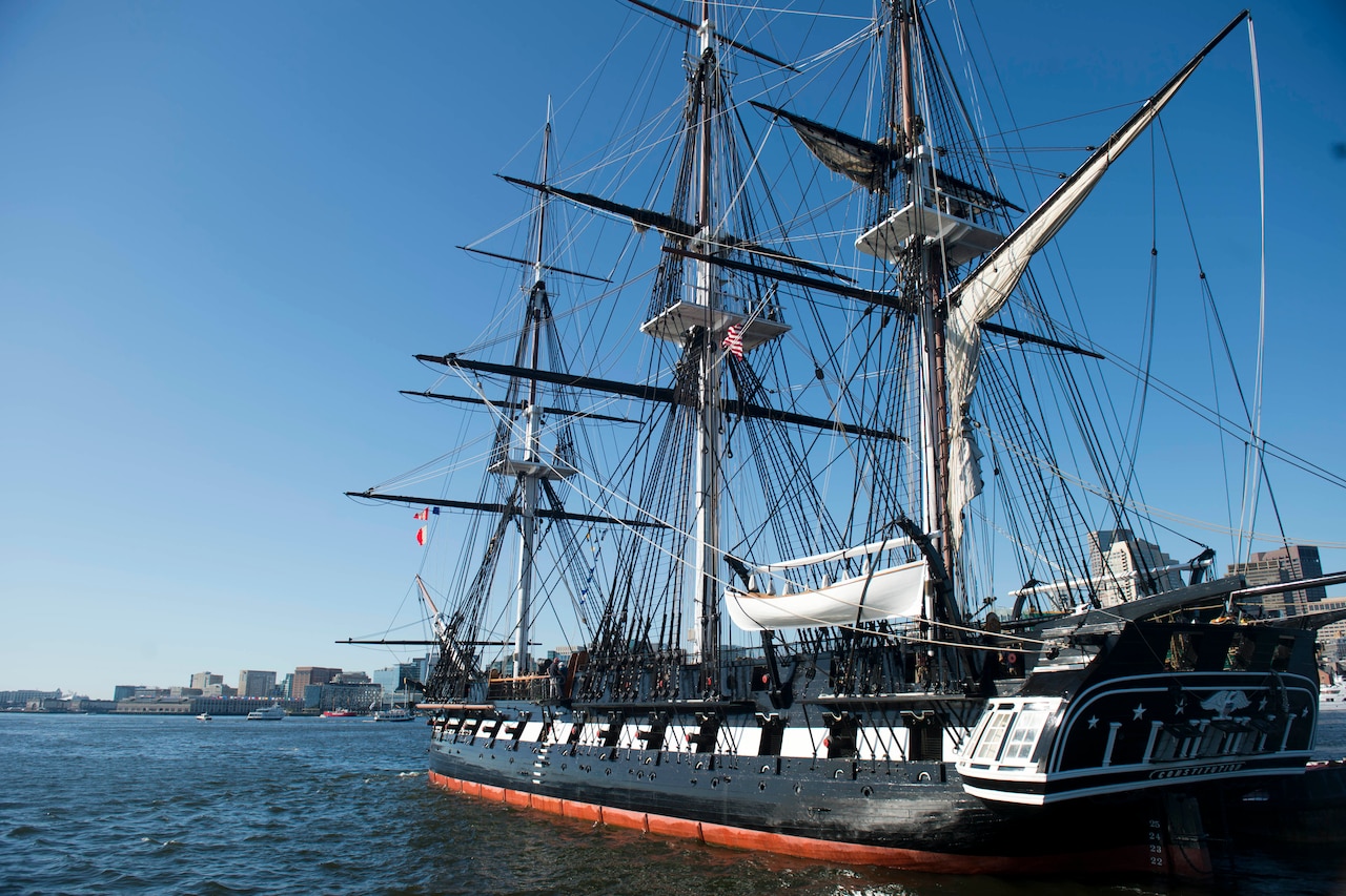 A three-mast wooden ship sails in a harbor.