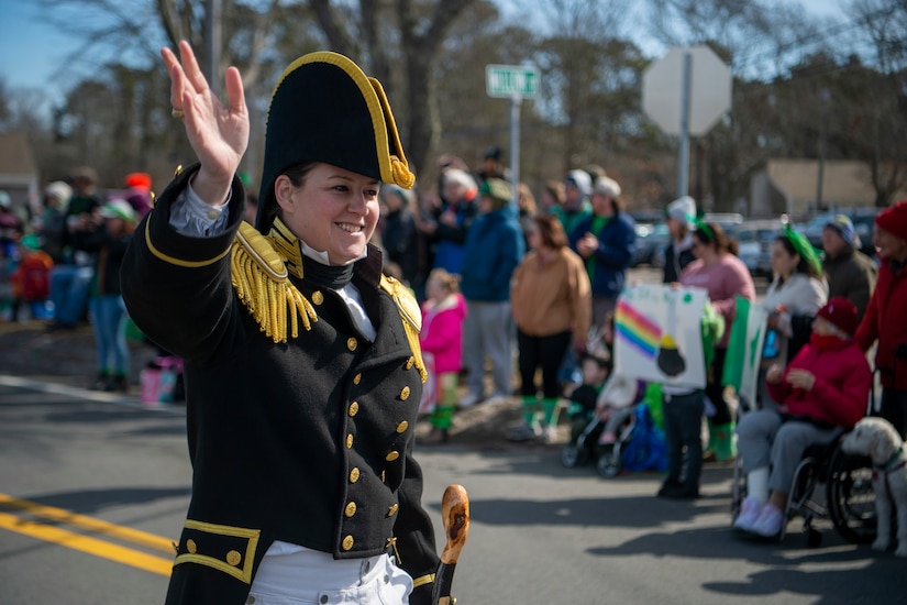 A woman in 1812-era Navy garb and cap waves while walking past a crowd.