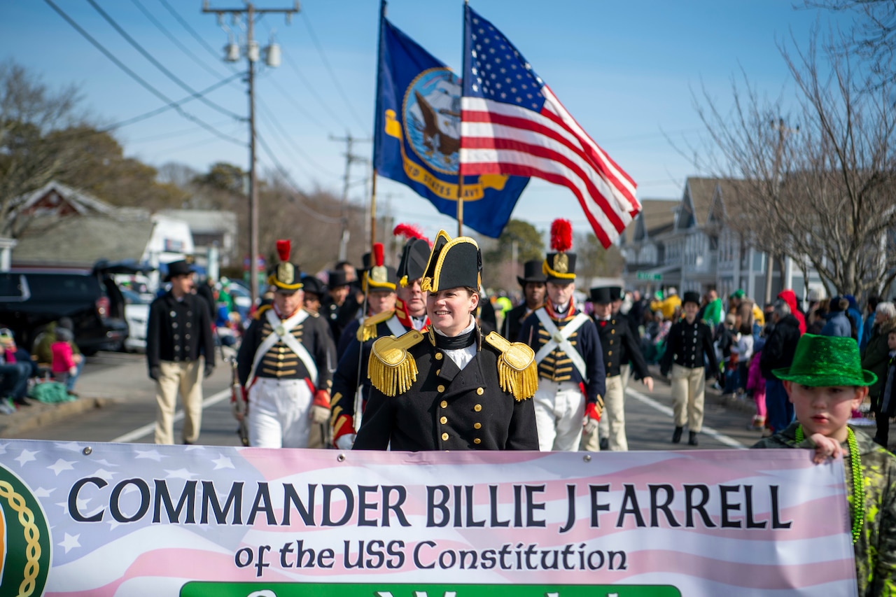 A woman in 1812-era Navy garb walks behind a large banner with her name on it. Behind her, other sailors march in formation while carrying the U.S. and Navy flag.