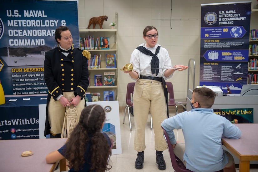 A sailor presents an 1812-era sword to a classroom of schoolchildren while another sailor stands by beside her.
